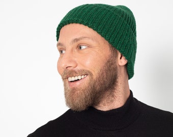 Puede incluir: Un hombre con una boina de punto verde y un jersey de cuello alto negro. Está sonriendo y mirando hacia la derecha.