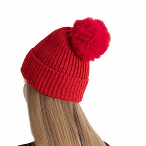 Puede incluir: Un gorro de punto rojo vibrante con un pompón a juego. El gorro es acanalado y parece estar hecho de un material suave y cálido. El gorro lo lleva una persona con pelo largo y rubio liso, sobre un fondo blanco.