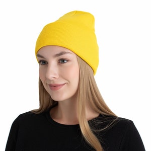 Könnte beinhalten: Eine leuchtend gelbe Beanie-Mütze. Die Mütze besteht aus einem gerippten Strickmaterial und wird auf dem Kopf getragen. Die Person auf dem Bild trägt ein schwarzes Langarmshirt. Der Hintergrund ist weiß.