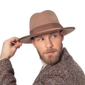 Könnte beinhalten: Ein Mann trägt einen braunen Fedora-Hut und eine braune Jacke mit blauem Muster. Er hält den Hut mit der rechten Hand am Rand fest und schaut nach rechts.