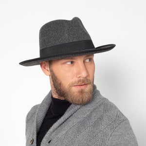 Può includere: Un uomo che indossa un fedora grigio con una fascia nera e un cardigan di maglia grigia con un dolcevita nero sotto.