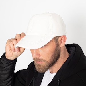May include: A white leather baseball cap with a curved brim. The cap is being held by a person wearing a black hooded jacket.
