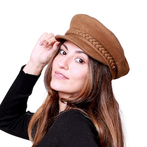 Peut inclure: Un chapeau de style gavroche marron avec une bande tressée et un petit bouton. Le chapeau est fait d'une matière douce et a un bord structuré. C'est un accessoire élégant.