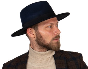 May include: A man wearing a dark blue fedora hat with a black band and a brown and black plaid jacket over a beige turtleneck sweater.