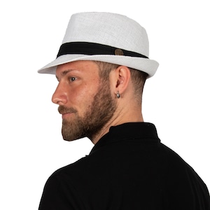 Puede incluir: Un sombrero fedora blanco con una banda negra y un pequeño botón marrón en la parte delantera. El sombrero está hecho de un material tejido y tiene un borde ligeramente curvado.