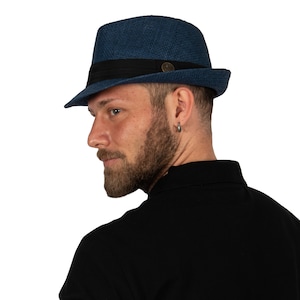 Könnte beinhalten: Ein blauer Fedora-Hut mit einem schwarzen Band und einem kleinen Metallknopf. Der Hut ist aus gewebtem Material gefertigt und hat einen leicht gebogenen Rand.