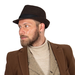 Puede incluir: Un hombre con un sombrero fedora marrón con una banda negra y un abrigo marrón sobre un jersey de cuello alto beige.