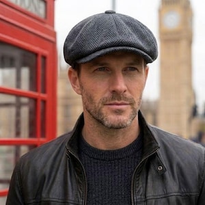 May include: A gray herringbone pattern newsboy cap. The cap is worn by a person wearing a black leather jacket and a dark blue sweater. The background includes a red telephone booth and a building with a clock tower.
