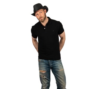 K&ouml;nnte beinhalten: Ein Mann tr&auml;gt ein schwarzes Poloshirt und eine blaue Jeans mit Destroyed-Details. Er tr&auml;gt einen schwarzen Cowboyhut.