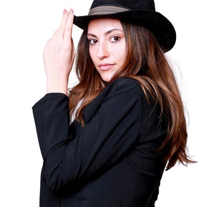 May include: A woman wearing a black blazer and a black fedora with a brown band. She is looking at the camera and has her hand on the brim of her hat.
