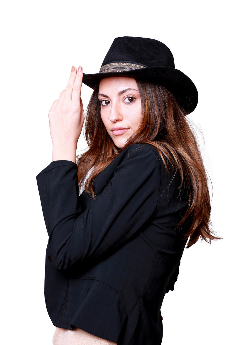 May include: A woman wearing a black blazer and a black fedora with a brown band. She is looking at the camera and has her hand on the brim of her hat.