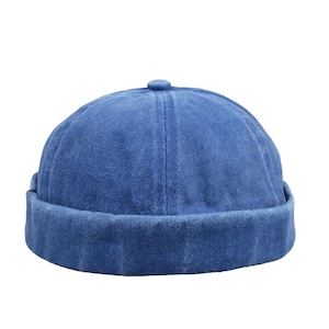 Puede incluir: Una gorra de calavera de mezclilla azul con un borde enrollado. El sombrero tiene una corona redondeada y un pequeño detalle de botón en la parte superior. La gorra está hecha de una tela suave y texturizada.