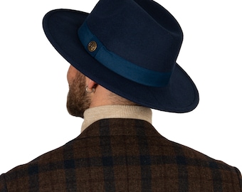 May include: A dark blue fedora hat with a dark blue ribbon and a gold button. The hat is worn by a person wearing a brown and black plaid jacket.