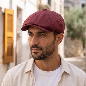 Pode incluir: Um boné estilo boina em tom bordô profundo com uma coroa estruturada, uma aba curta e curva, e um pequeno botão revestido de tecido no centro do topo. O boné é feito de tecido de cor sólida com detalhes de costura visíveis nos painéis.