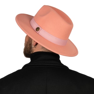 May include: A pink fedora hat with a pink ribbon and a gold button. The hat is made of felt and has a wide brim.