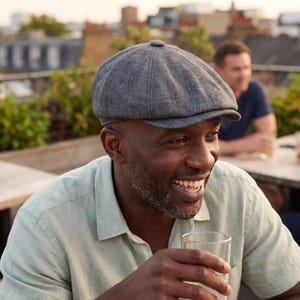 May include: A charcoal grey flat cap with a structured crown and a short, stiff brim. The cap features a textured fabric finish and a small button detail at the top centre, providing a classic silhouette that sits snugly on the head.