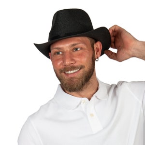 K&ouml;nnte beinhalten: Ein Mann mit einem schwarzen Fedora-Hut und einem wei&szlig;en Poloshirt. Er l&auml;chelt und schaut zur Seite.