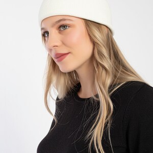 K&ouml;nnte beinhalten: Eine wei&szlig;e Strickm&uuml;tze mit gerippter Struktur. Die M&uuml;tze wird von einer Person mit langen blonden Haaren und einem schwarzen Pullover getragen.