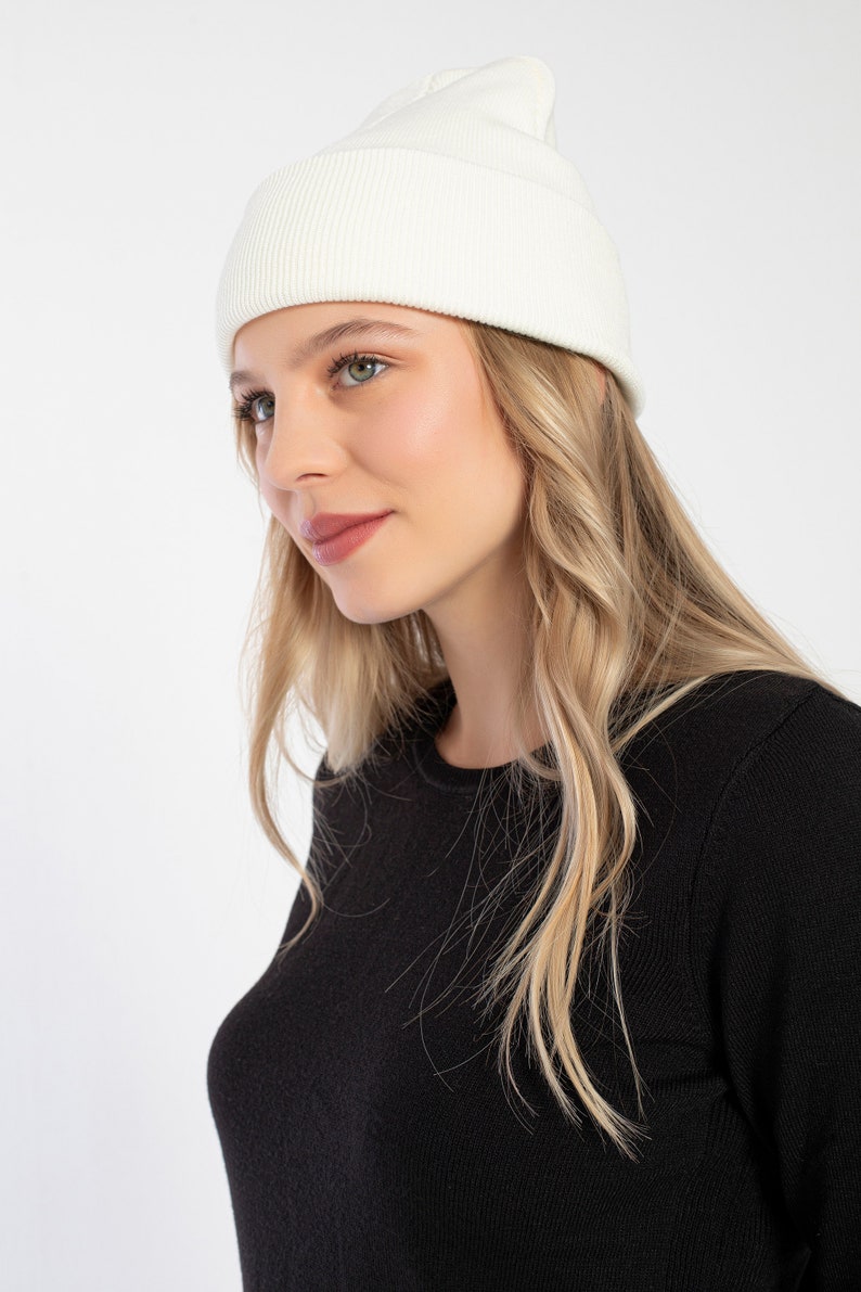 K&ouml;nnte beinhalten: Eine wei&szlig;e Strickm&uuml;tze mit gerippter Struktur. Die M&uuml;tze wird von einer Person mit langen blonden Haaren und einem schwarzen Pullover getragen.