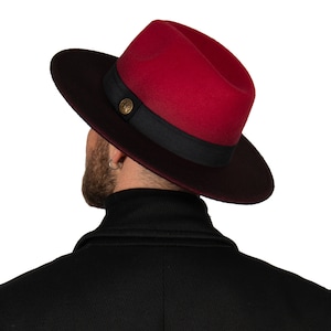 Puede incluir: Un sombrero fedora rojo y negro con una banda negra y un botón dorado.