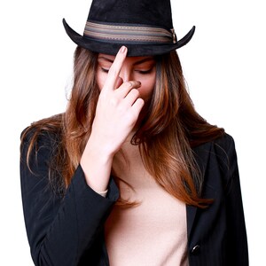 May include: A woman wearing a black blazer and a brown fedora with a striped band. She is looking down and touching the brim of her hat with her finger.