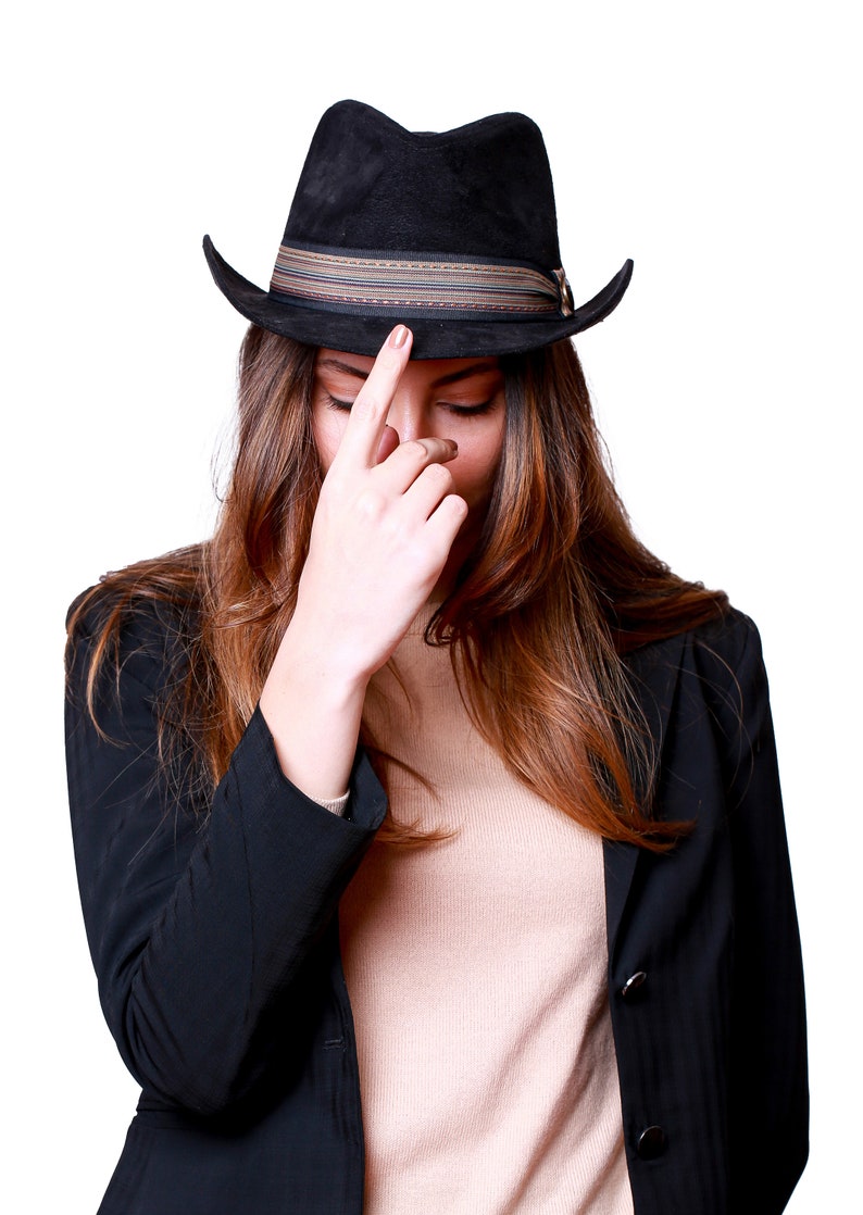 May include: A woman wearing a black blazer and a brown fedora with a striped band. She is looking down and touching the brim of her hat with her finger.