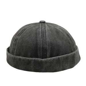 Può includere: Un berretto senza tesa grigio scuro con una corona arrotondata e un bordo ripiegato. Il cappello ha un aspetto strutturato e un piccolo dettaglio a bottone sulla parte superiore. È un accessorio casual.