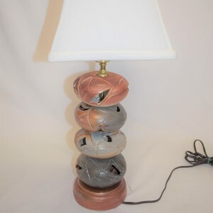 May include: A decorative table lamp with a white lampshade. The lamp base is composed of stacked, spherical, ceramic elements in shades of copper, gray, and bronze. The base has cut-out leaf designs and a round base.