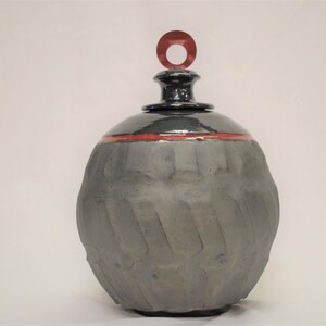 May include: A black ceramic jar with a red rim and a red ring on the lid. The jar has a textured surface and a round shape.