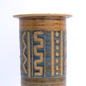 May include: A brown ceramic vase with a geometric design in blue and brown. The vase has a wide mouth and a rounded base.