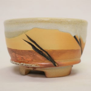 May include: A small, ceramic planter with a light brown, orange, and white glaze. The planter has three small feet and black brushstrokes on the side.