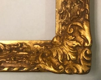 May include: A gold-colored picture frame with an ornate, detailed design. The frame has a raised, textured border with intricate carvings.