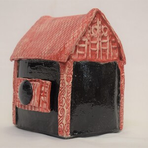 May include: A small red and black ceramic house with a detailed design. The house has a red roof and black walls with white accents. The house has a small door with a black knob.