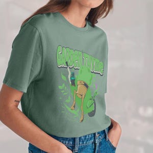 May include: Sage green t-shirt with the text "GARDEN THYME" in a green, outlined font. The graphic features a cartoon plant pot riding a green scooter, with green leaves and smoke-like elements.