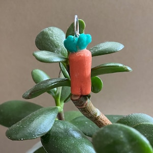 Puede incluir: Un charm de zanahoria hecho a mano con un lazo plateado. La zanahoria es naranja con hojas verdes. El charm cuelga de una planta verde con hojas ovaladas.