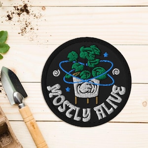 May include: A black embroidered patch with a green plant in a white pot. The plant is surrounded by a blue circle with stars. The text "Mostly Alive" is written in a white, cursive font around the circle.