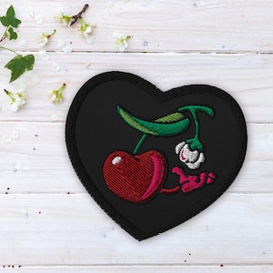 Puede incluir: Parche negro en forma de corazón con una cereza bordada y un tallo. La cereza es roja con un tallo y hojas verdes. La cereza tiene una flor blanca en el tallo.