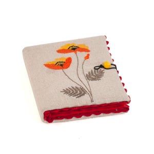 May include: A beige fabric journal with an embroidered floral design featuring orange and yellow poppies. The journal has a red scalloped edge and a yellow button closure. The cover is made of a textured fabric.