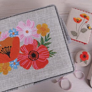 K&ouml;nnte beinhalten: Eine graue N&auml;hbox mit floraler Stickerei mit roten Mohnblumen, rosa und gelben Blumen. Weitere N&auml;haccessoires sind ein Nadelbuch, eine Schere, ein Ma&szlig;band und Garnrollen.