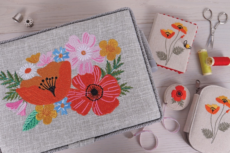 K&ouml;nnte beinhalten: Eine graue N&auml;hbox mit floraler Stickerei mit roten Mohnblumen, rosa und gelben Blumen. Weitere N&auml;haccessoires sind ein Nadelbuch, eine Schere, ein Ma&szlig;band und Garnrollen.