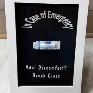 Könnte beinhalten: Ein weiß gerahmtes Kunstwerk mit schwarzem Hintergrund. Weißer Text liest sich: „In Case of Emergency“ und „Anal Discomfort? Break Glass“. Ein Zäpfchen ist abgebildet.