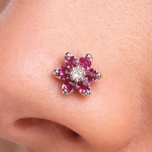 May include: A nose stud with a flower design. The stud has a silver setting with six pink gemstones around a central clear gemstone. The jewellery is worn on the nose.