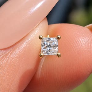 Puede incluir: Un pendiente de diamante de talla cuadrada con engaste de oro. El diamante transparente y brillante está sujeto por cuatro pequeñas garras de oro. El pendiente es una toma de primer plano, destacando los detalles de la joya.