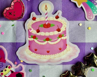 Peut inclure: Un autocollant rose représentant un gâteau d'anniversaire avec du glaçage blanc, des cerises rouges et une bougie allumée. Le gâteau a une rose sur le dessus et est décoré de cœurs. Autres autocollants : un cœur rose, un arc-en-ciel et des étoiles dorées.