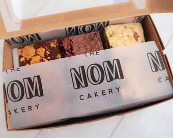 May include: A box of assorted brownies from The Nom Cakery. The box is brown with a clear plastic window on top. The brownies are arranged in the box with a white paper liner that has the company's logo printed on it.