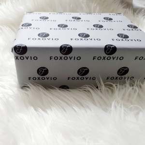 Könnte beinhalten: Eine weiße Geschenkbox mit einem sich wiederholenden Muster aus schwarzen Kreisen, die das Wort "FOXOVIO" in einer stilisierten Schrift enthalten.