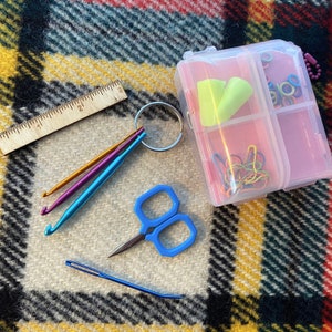 May include: A clear plastic storage box with multiple compartments containing various craft supplies, including colorful yarn, earplugs, and a pair of small scissors. The box is sitting on a plaid fabric surface.
