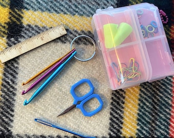May include: A clear plastic storage box with multiple compartments containing various craft supplies, including colorful yarn, earplugs, and a pair of small scissors. The box is sitting on a plaid fabric surface.