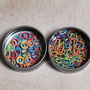 May include: Two round metal tins filled with colorful craft supplies. One tin contains a variety of colorful plastic rings, the other contains a variety of colorful safety pins.