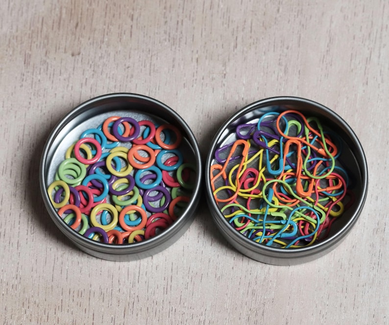 May include: Two round metal tins filled with colorful craft supplies. One tin contains a variety of colorful plastic rings, the other contains a variety of colorful safety pins.